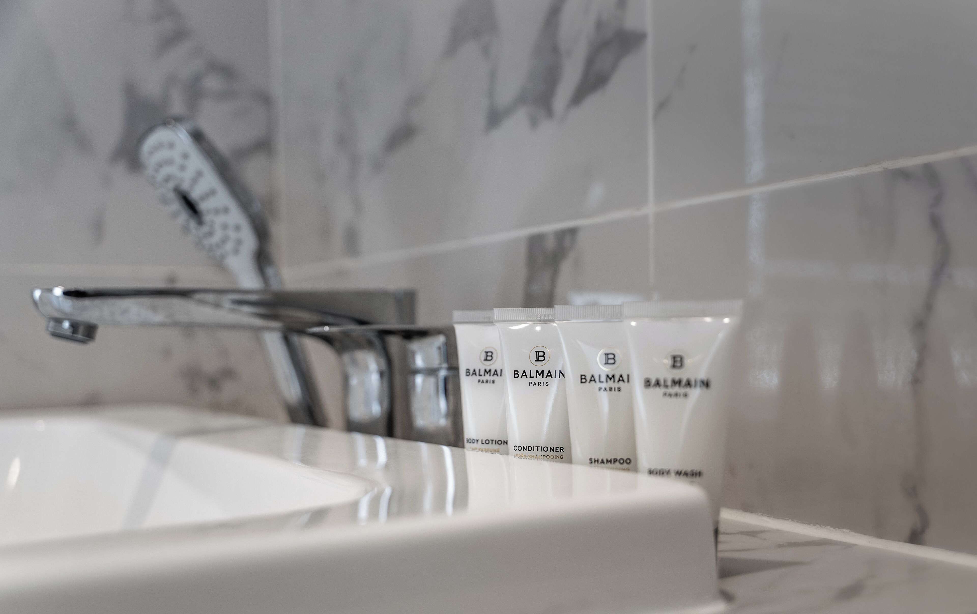 Un baño con un lavabo blanco, un grifo moderno y varios productos de cuidado de la piel Balmain Paris.