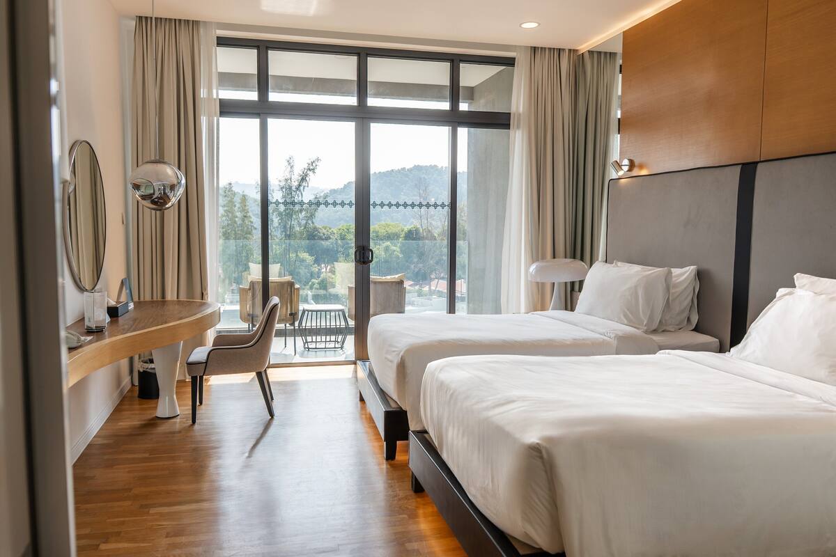 A hotel room with two beds, a desk, a chair, and a large window offering a view of the outdoors.