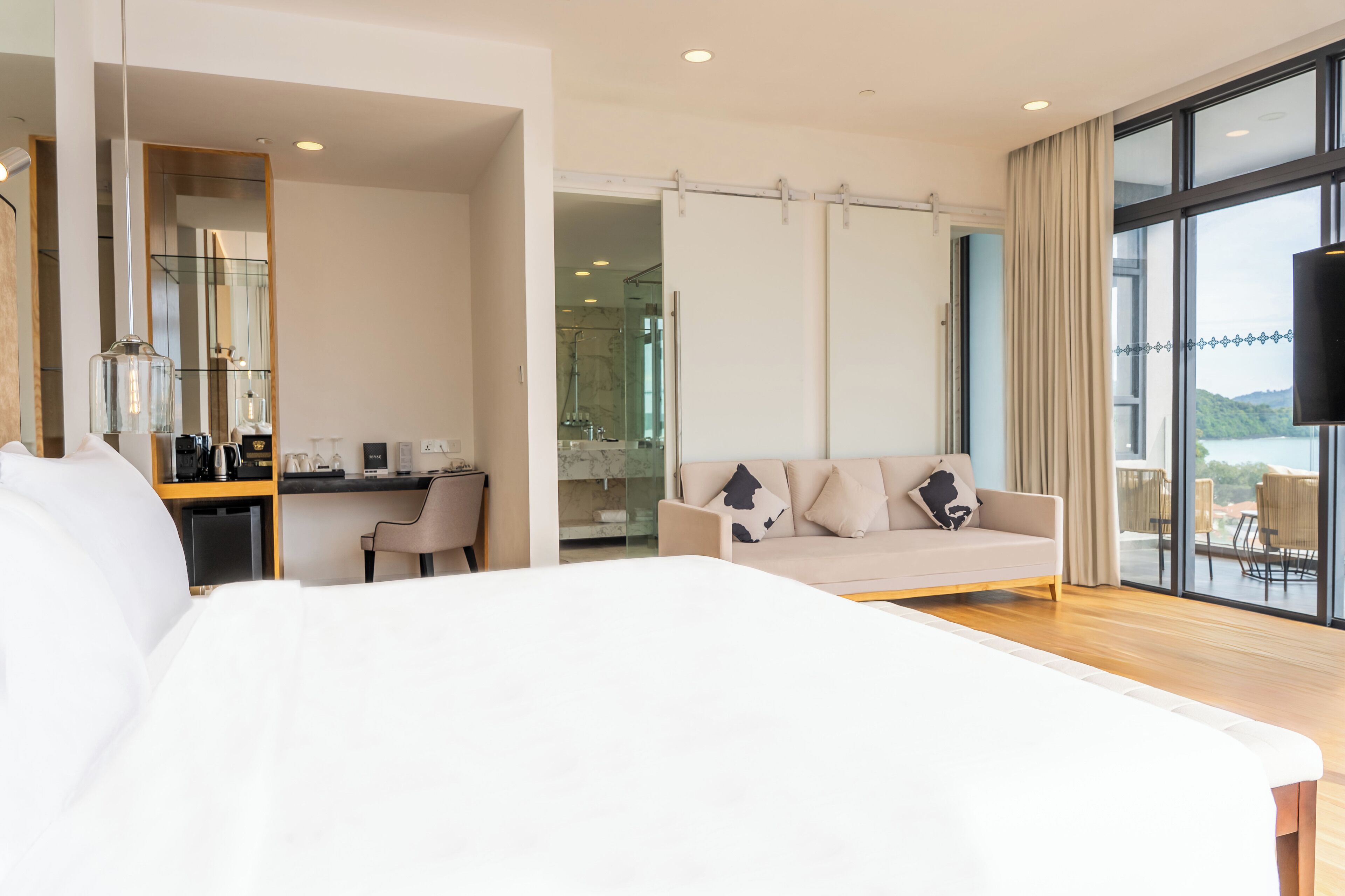 A modern hotel room with a large bed, a desk, a sofa, and a balcony with a view.