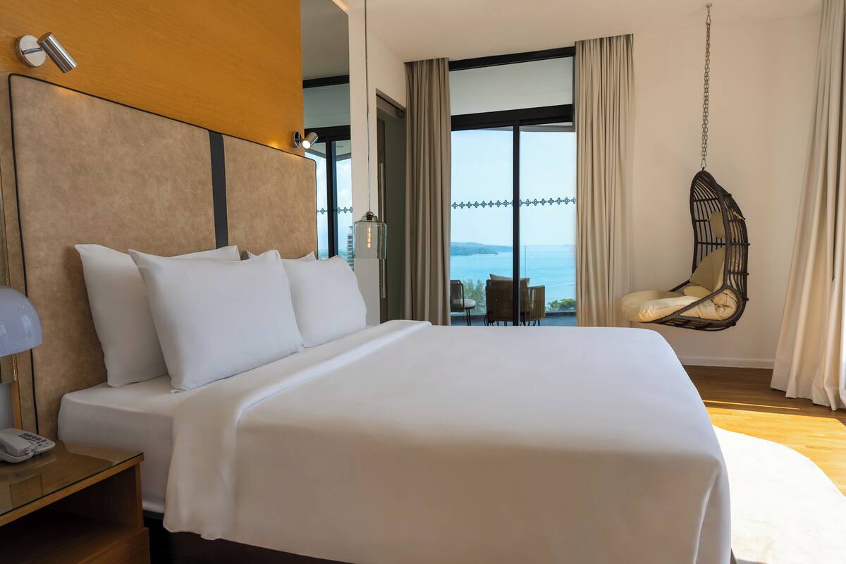 A large bed with white linens, a bedside table with a lamp and phone, a hanging chair, and a view of the sea through large windows.