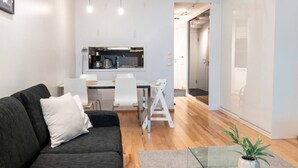 Ein modernes Wohnzimmer mit einem dunklen Sofa, einem Esstisch mit Stühlen, einer Küchenzeile und einer Schiebetür zu einem weiteren Raum.