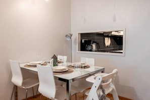 Uma área de refeições com uma mesa posta para quatro pessoas, cadeiras brancas, uma cafeteira e uma janela com utensílios de cozinha à mostra.