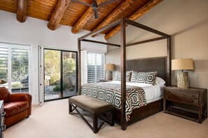 A four-poster bed with a patterned bedspread, a wooden nightstand with a lamp, a bench, and a leather sofa in a room with a wooden ceiling and large windows.