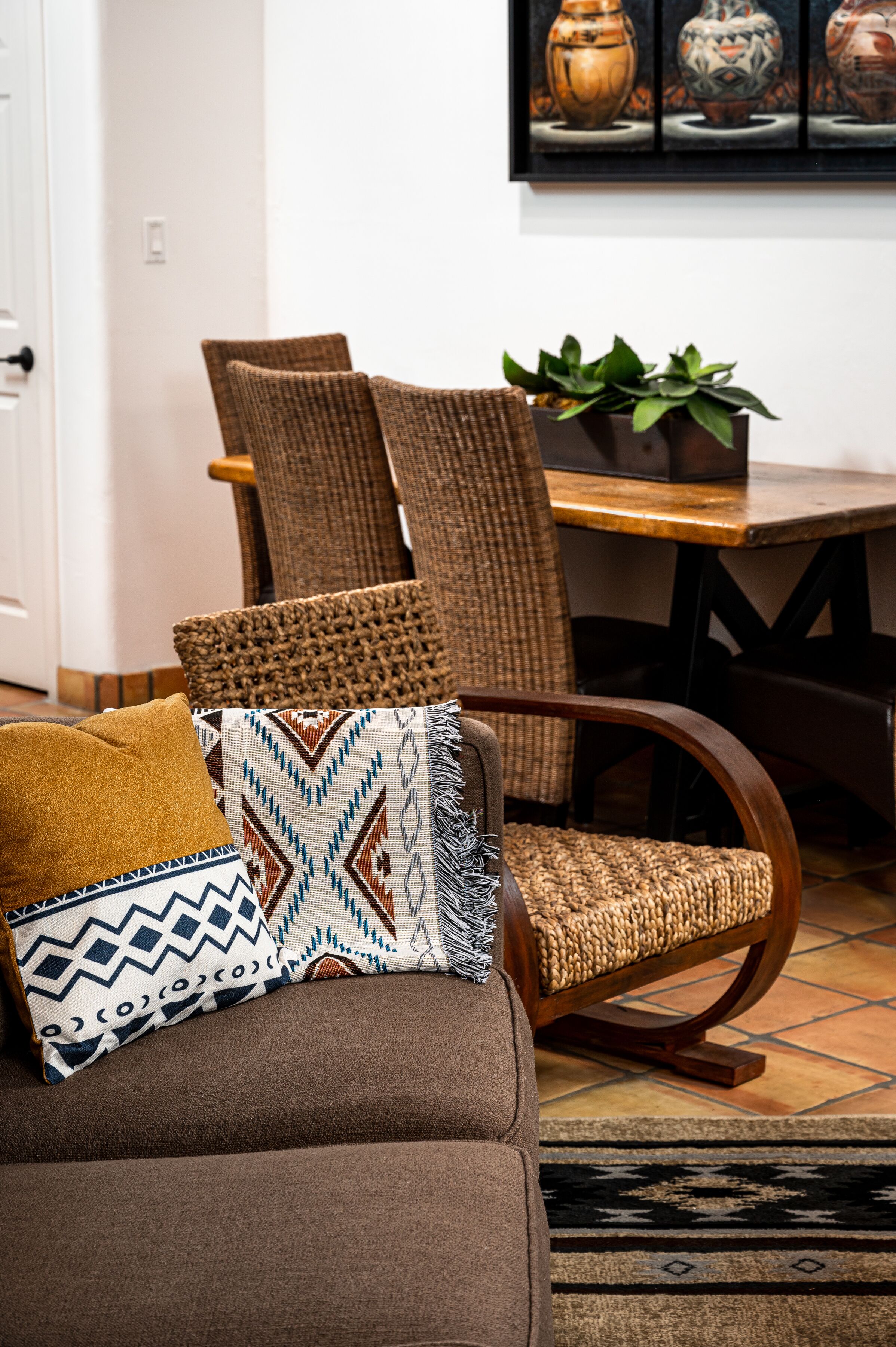 A brown sofa with patterned cushions, a wicker chair, and a wooden table with a plant.