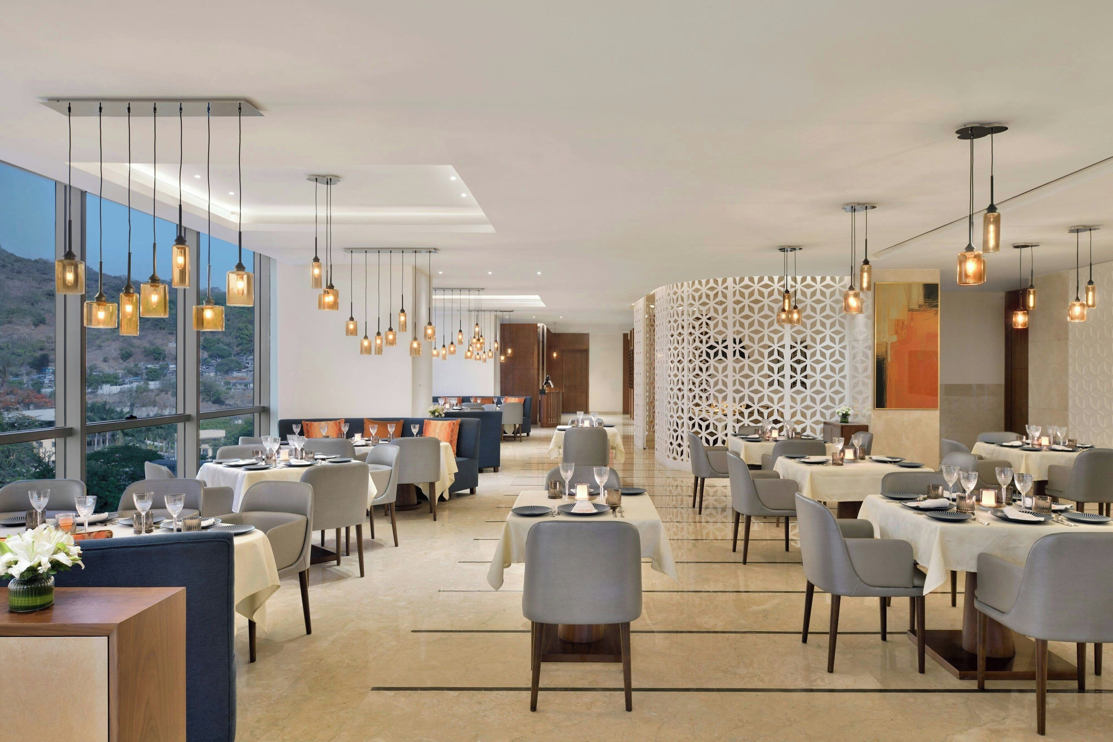 A modern dining area with tables set for service, large windows offering a view, and hanging pendant lights.