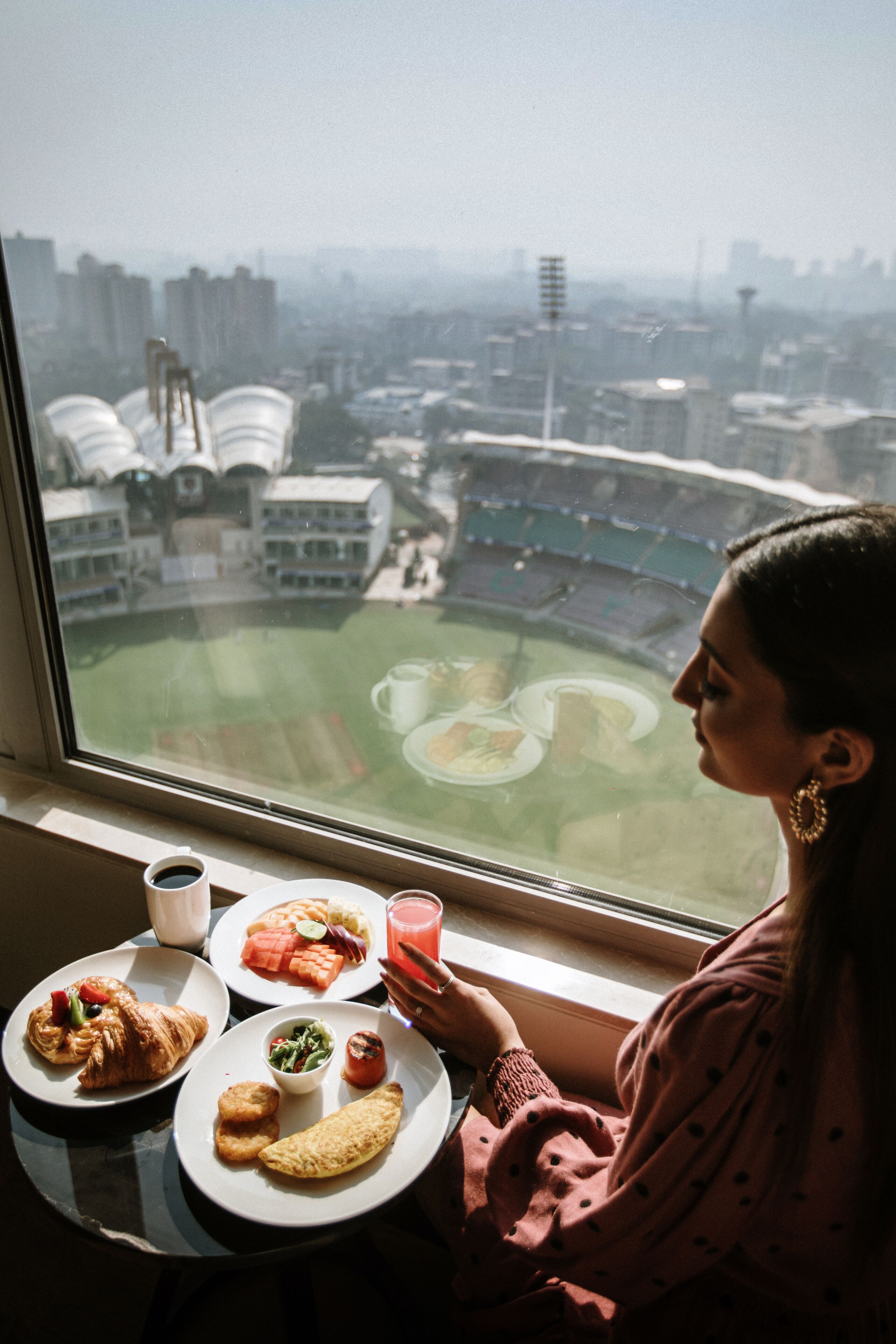 A person seated by a window overlooking a stadium with a meal on a table.