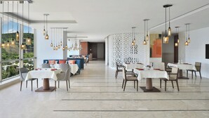 Un comedor moderno con varias mesas y sillas, lámparas colgantes y vistas a zonas verdes a través de amplios ventanales.