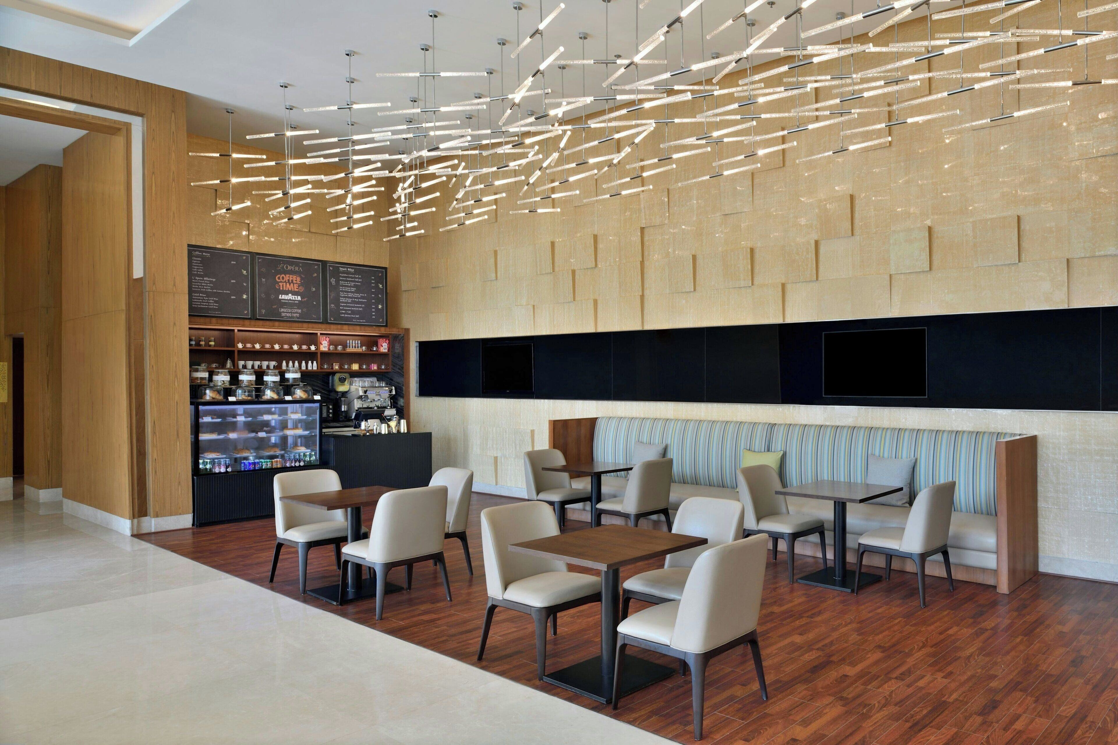 Una cafetería moderna con suelo de madera, sillas y mesas blancas. Se observa una barra con un menú y un expositor refrigerado.
