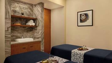 Un cuarto de spa con dos camillas de masaje, una pared de mármol, armarios de madera, un lavamanos y un cuadro abstracto enmarcado.