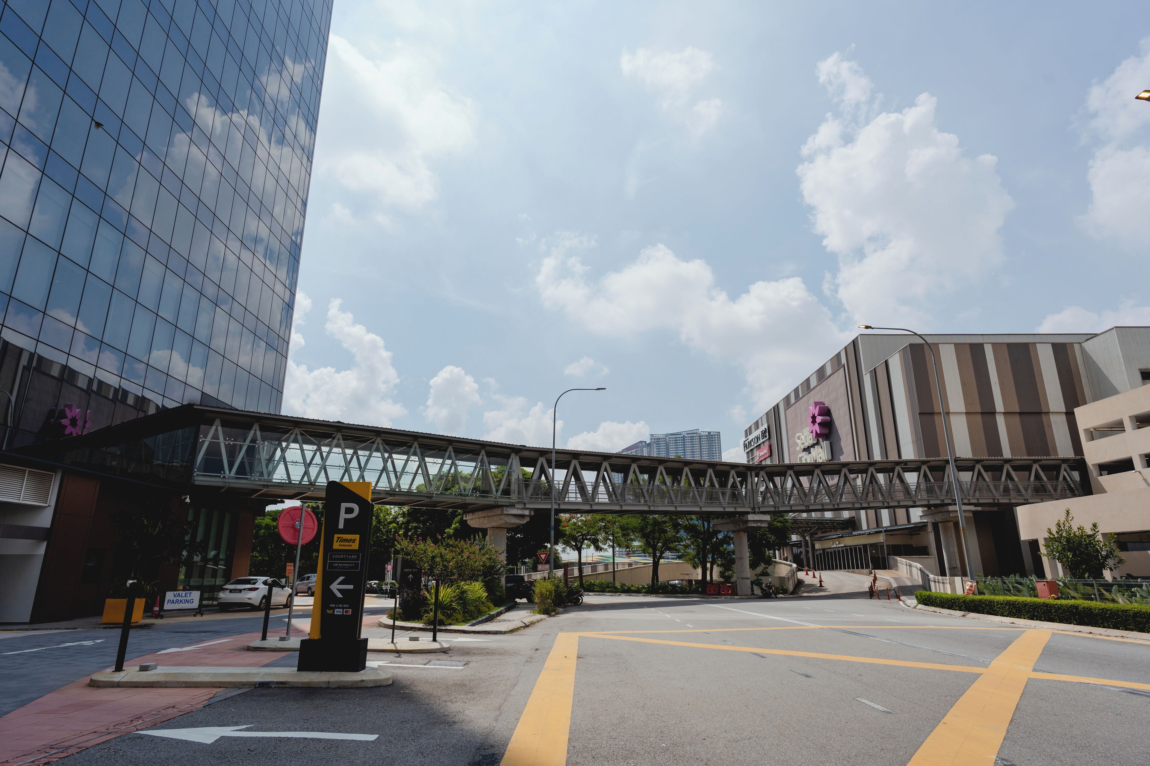 A modern urban intersection with a pedestrian bridge, a parking sign, and a building with reflective glass facade.