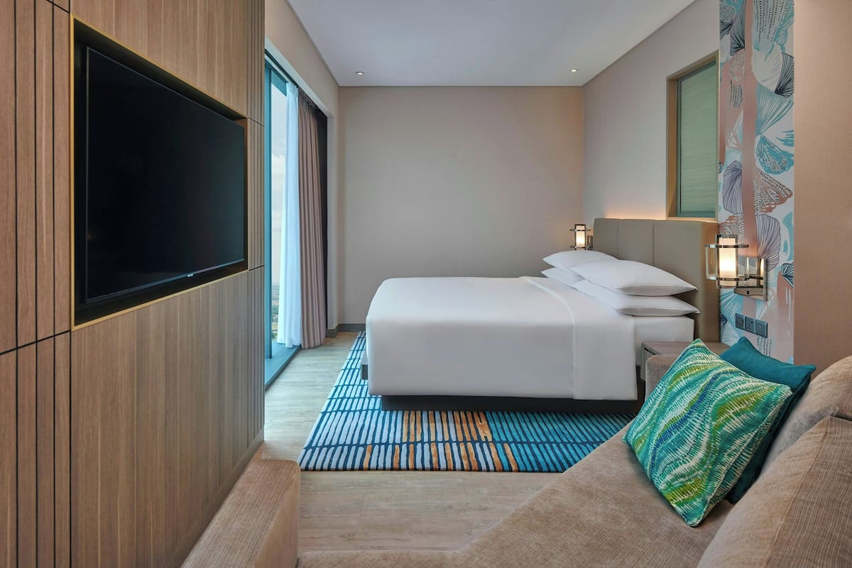 Habitación de hotel con una televisión de pantalla plana grande montada en la pared, una cama con ropa blanca, un tapete estampado y un sofá con un cojín decorativo.