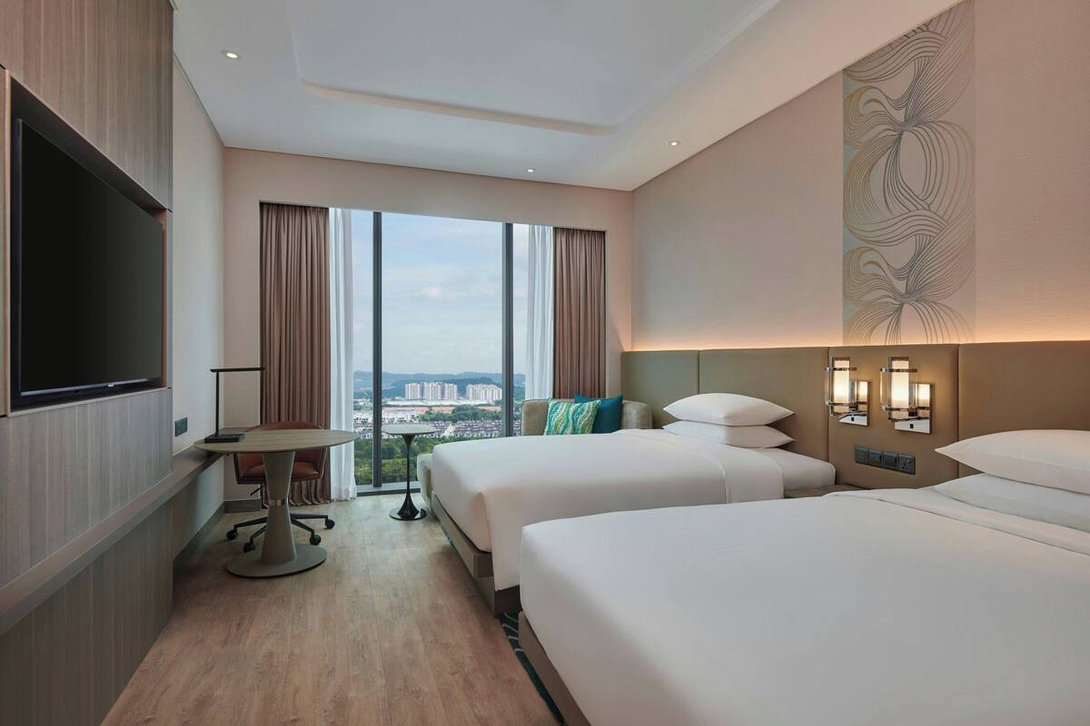 Habitación de hotel con dos camas, televisión, escritorio y ventana grande con vistas a la ciudad.