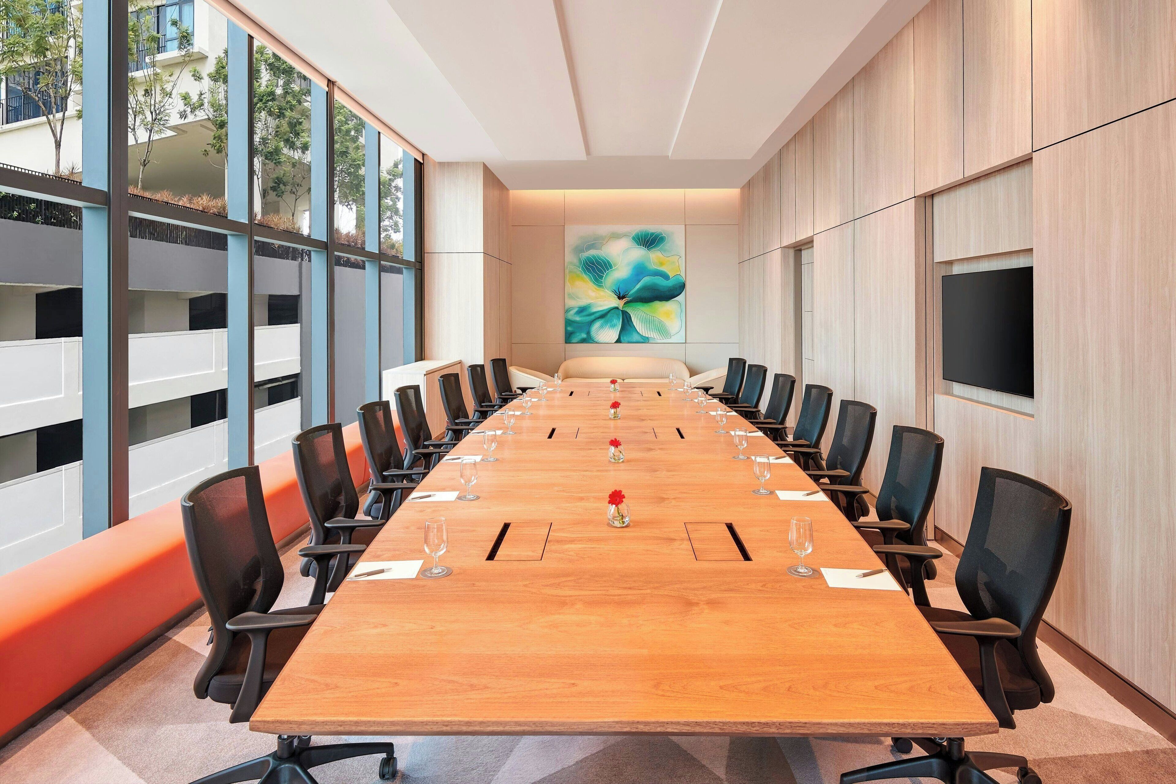 Une salle de conférence avec une longue table en bois, des chaises noires et une grande fenêtre donnant sur des immeubles et des arbres.