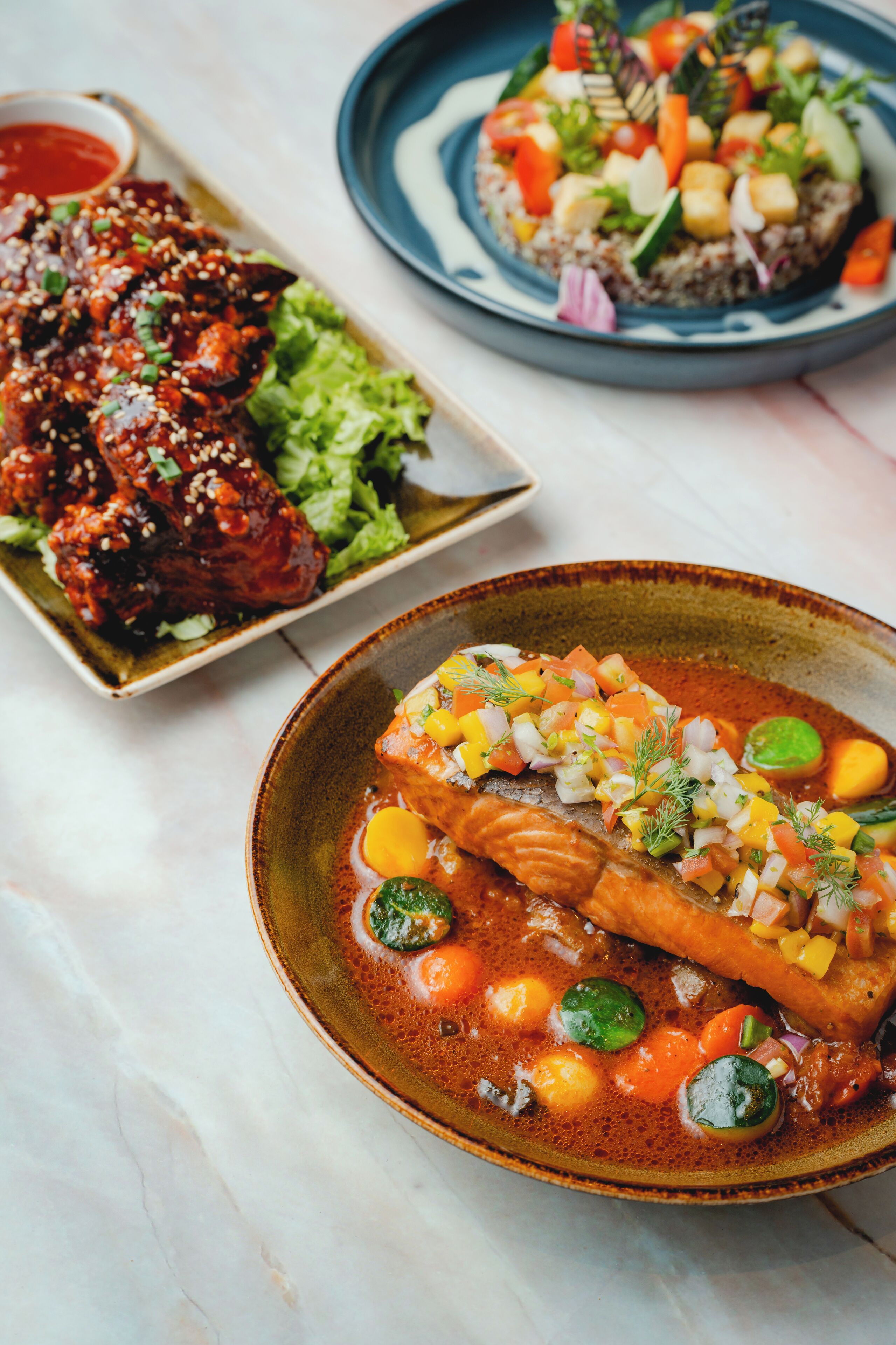 Diversos pratos, incluindo uma tigela de peixe com legumes, um prato de asas de frango e uma salada.
