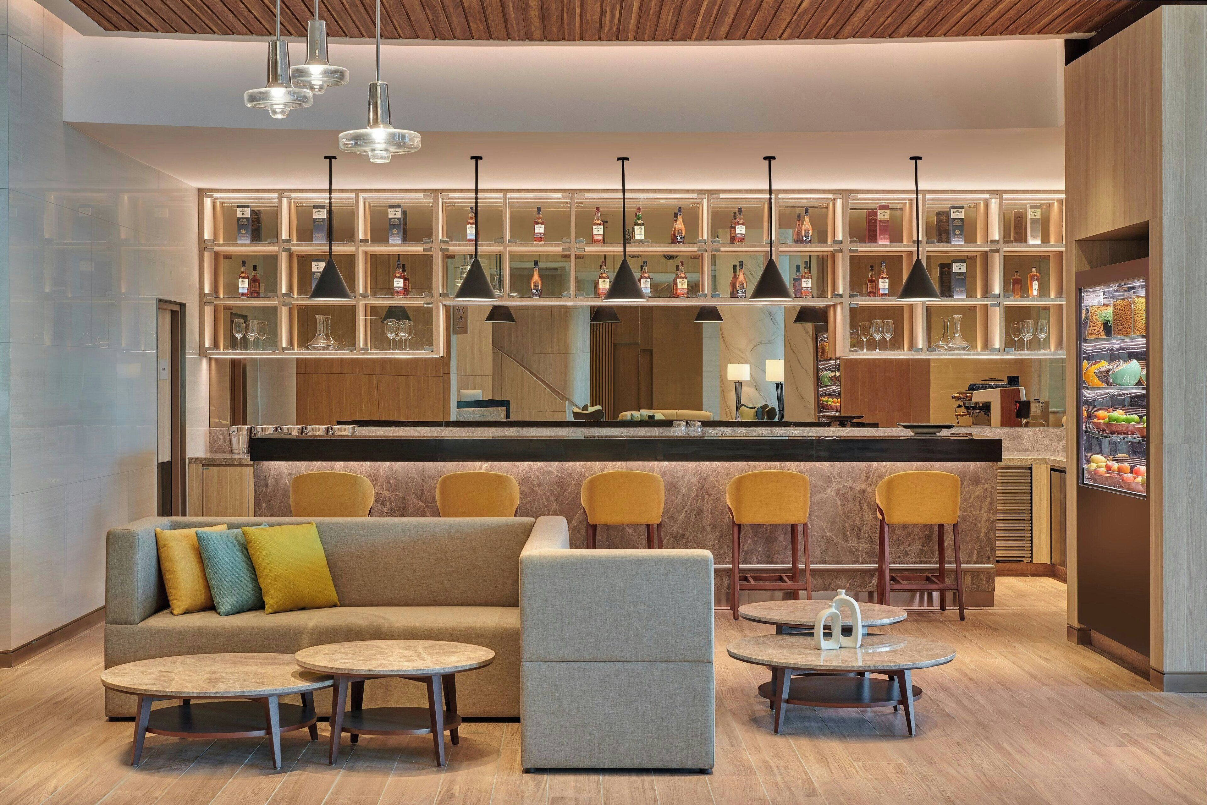 A modern bar area with a sofa, round tables, and bar stools. There are shelves with bottles and glasses, and a refrigerator with fruits.
