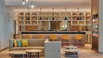 A modern bar area with a sofa, round tables, and bar stools. There are shelves with bottles and glasses, and a refrigerator with fruits.