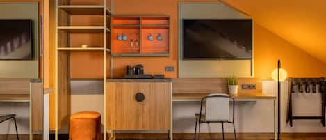 A modern hotel room with a loft bed, a desk with a computer, a chair, a small orange ottoman, a shelf with decorative items, and a large window.
