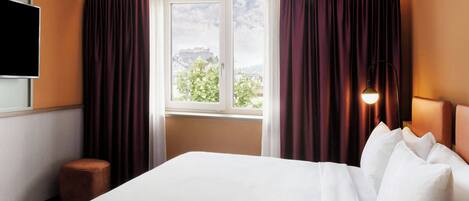 A hotel room with a large bed, a flat-screen TV mounted on the wall, a window with curtains, and a nightstand with a lamp.