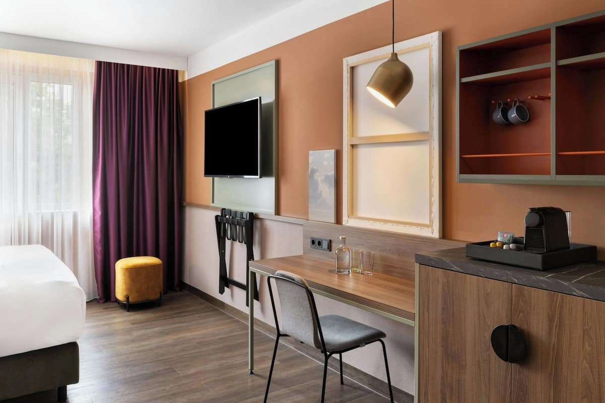 A hotel room with a bed, a desk, a chair, a TV, a coffee machine, and a shelf with cups.