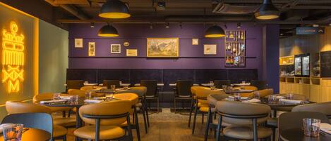 A restaurant with round tables and chairs, purple walls, and a neon sign.