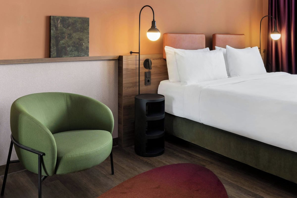 A hotel room with a bed, a green armchair, a side table, and a wall-mounted lamp.