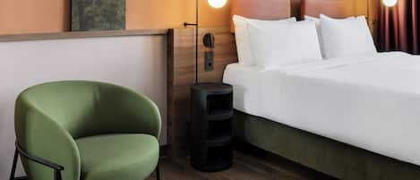 Habitación de hotel con una cama, un sillón verde, una mesita auxiliar y una lámpara de pared.
