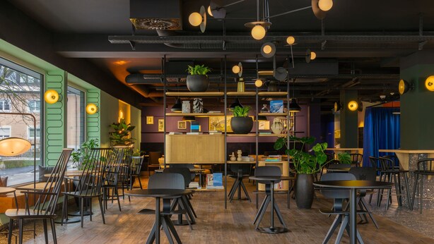 A modern cafe with wooden flooring, black chairs, and tables. There are hanging lights and shelves with plants and books. - June Six Salzburg, a Tribute Portfolio Hotel (Salzburg)