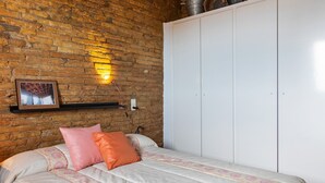 Makuuhuone, jossa on tiiliseinä, kuviollinen päiväpeite sängyllä, valkoinen vaatekaappi ja seinään kiinnitetty valaisin.