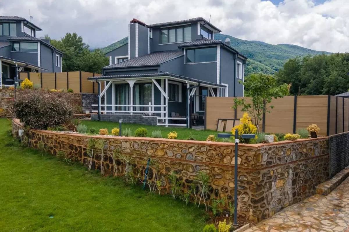 A modern two-story house with a stone retaining wall and a grassy yard.