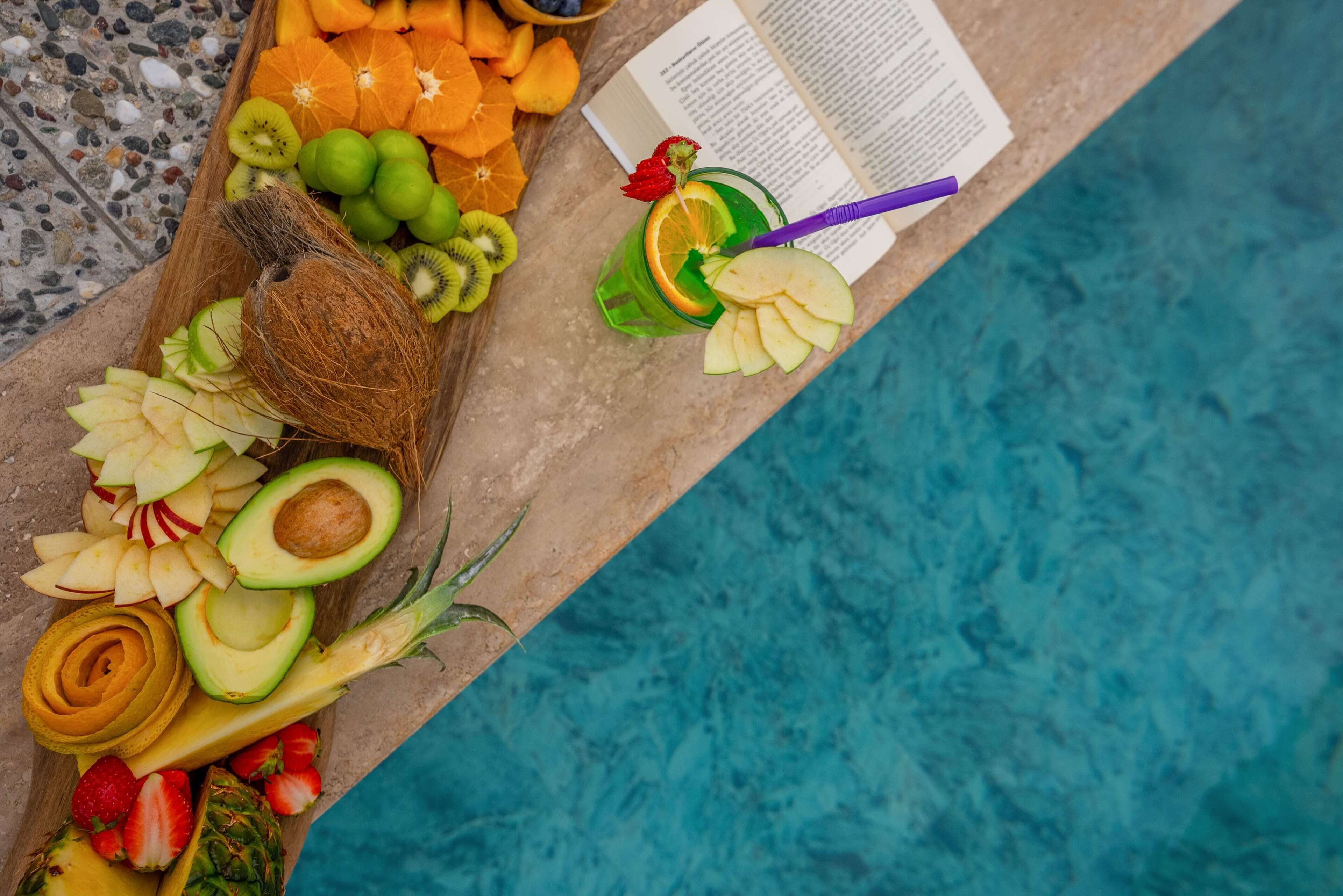 Frutas variadas y una bebida sobre una mesa junto a la piscina, con un libro abierto.