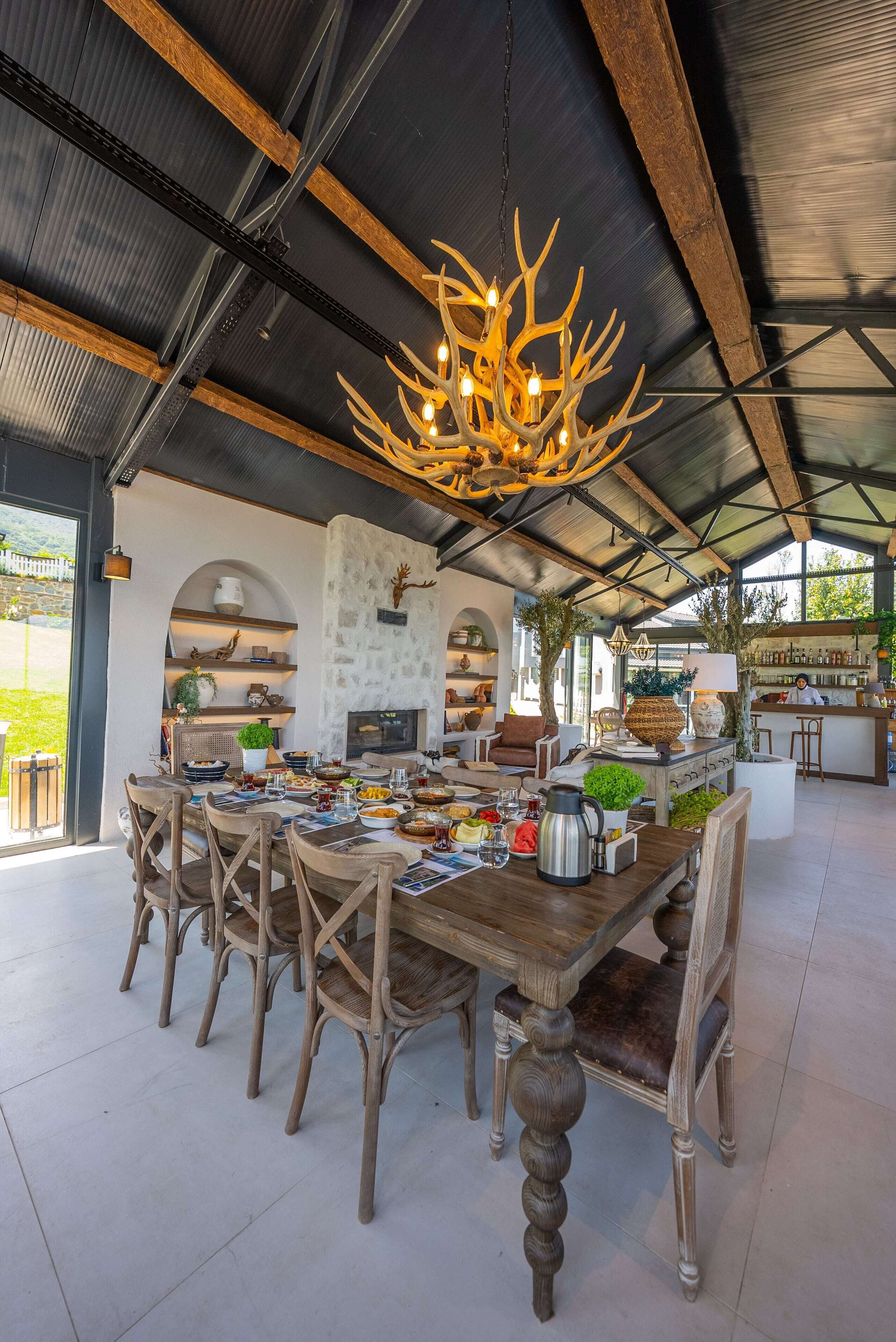 A spacious dining area with a large wooden table and chairs, a chandelier, and a kitchen in the background.
