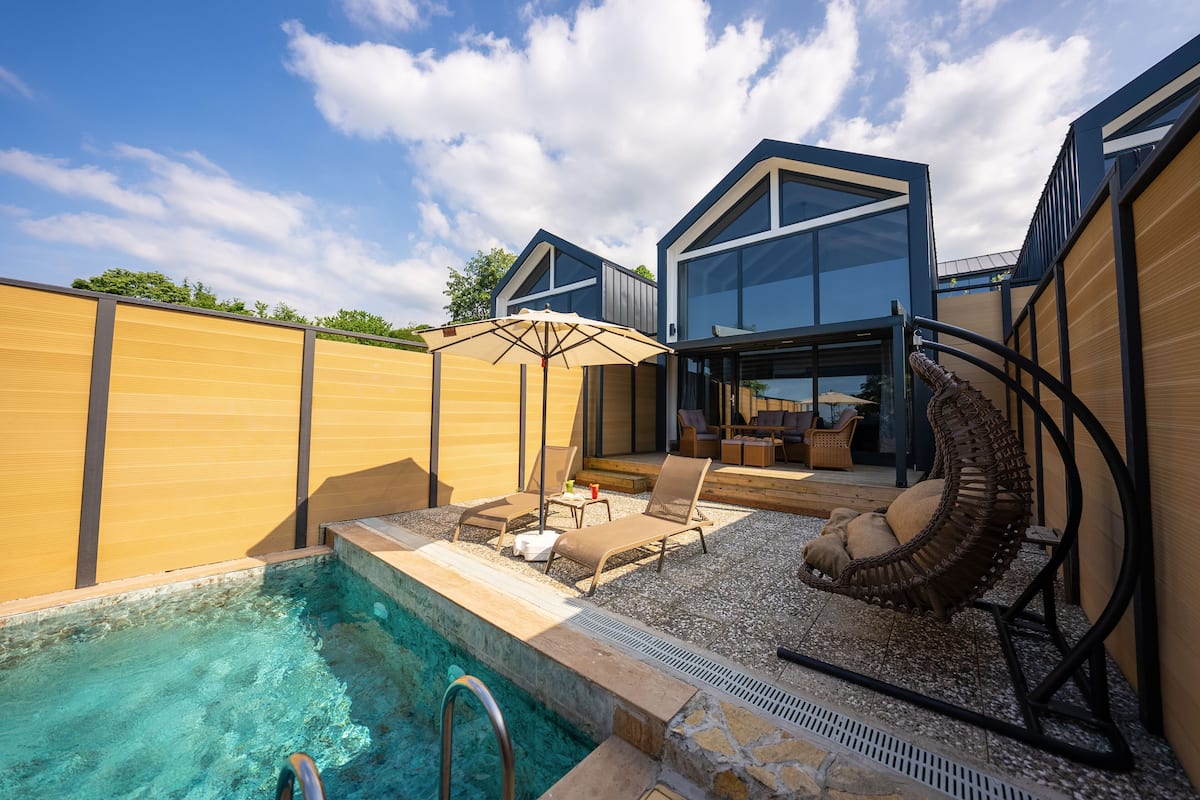 A modern outdoor pool area with lounge chairs and a wicker chair, surrounded by wooden fences and a clear blue sky.