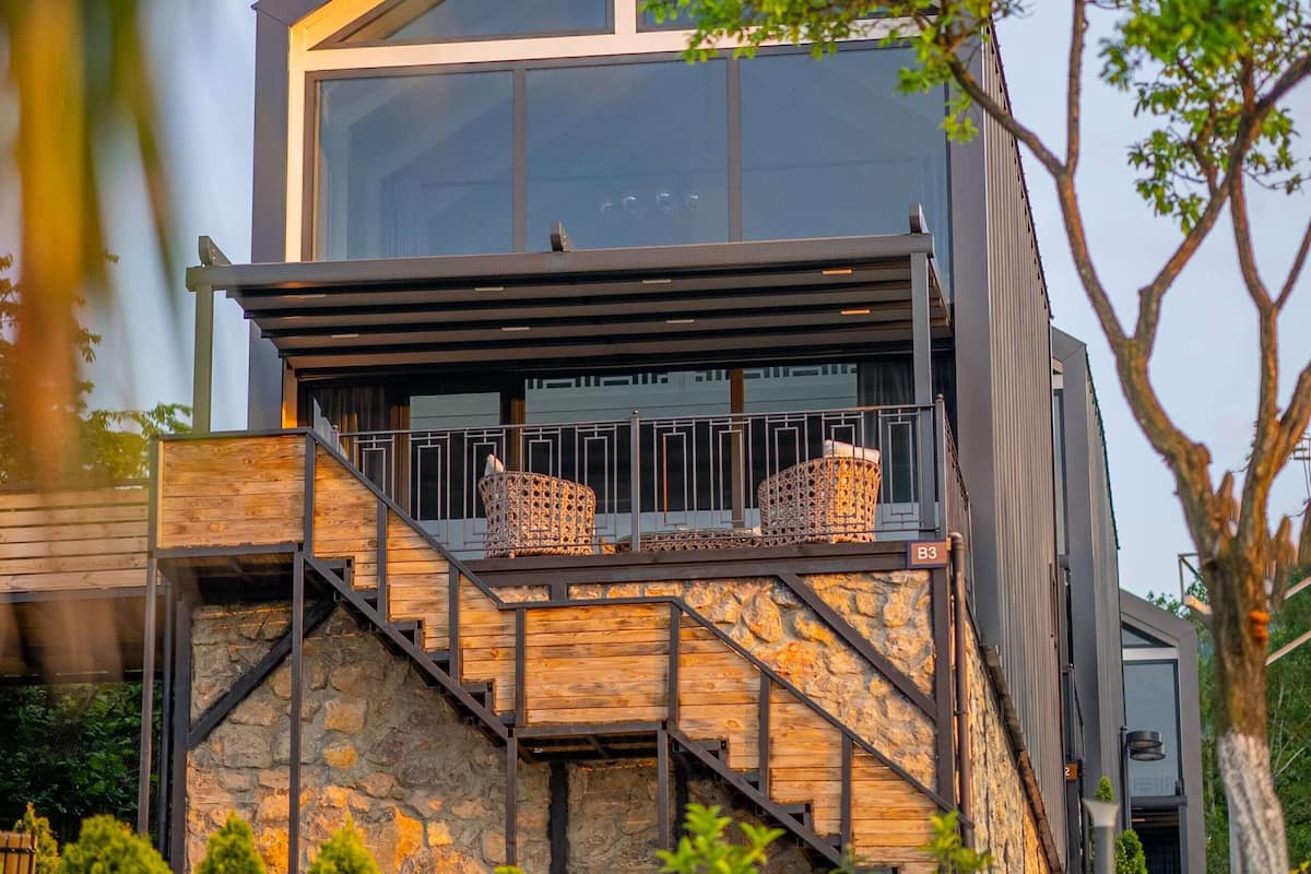 A modern two-story building with a stone facade, a balcony with wicker furniture, and a clear view of the sky.