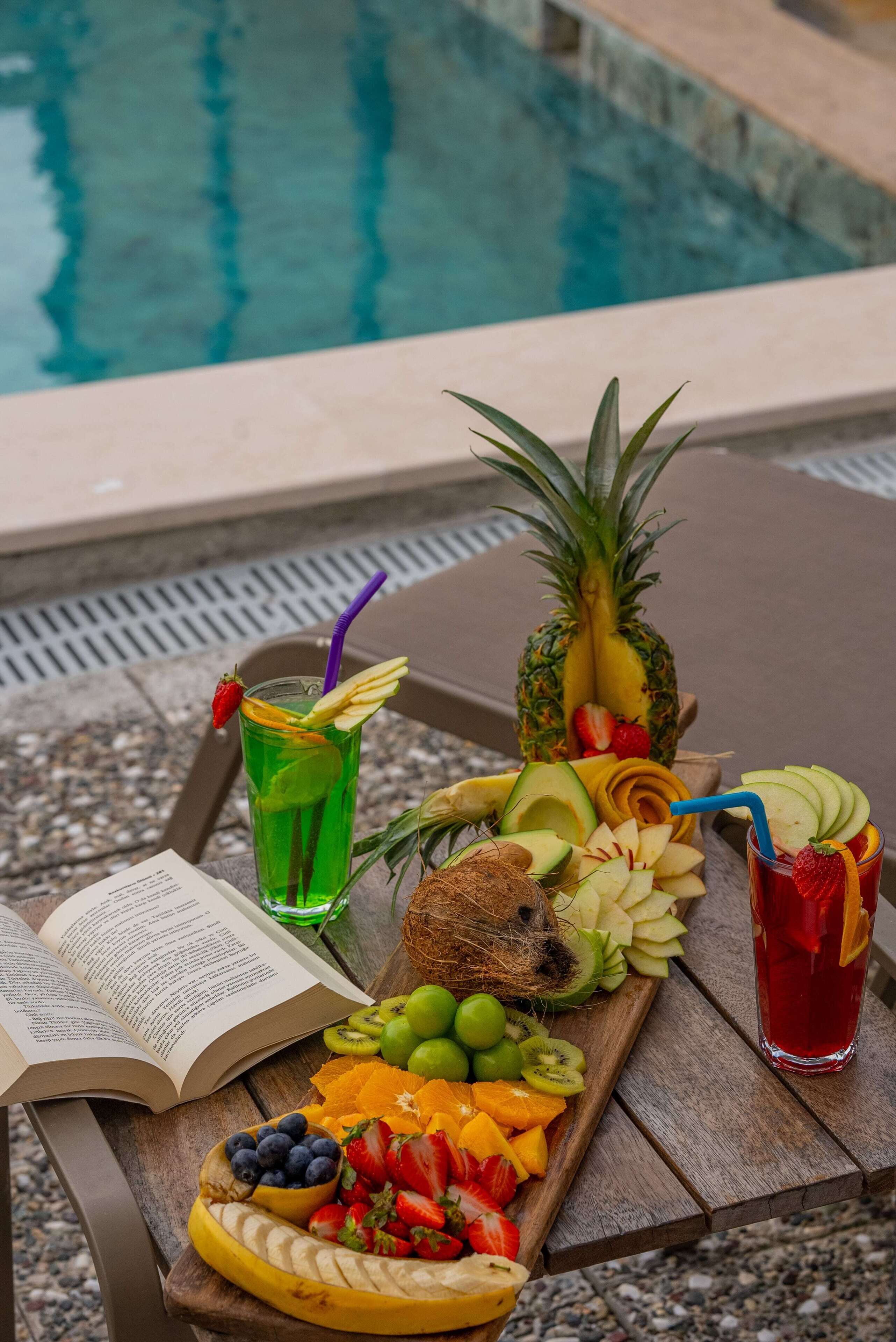 Un plateau en bois contenant une variété de fruits, une boisson et un livre ouvert, posé près d’une piscine.