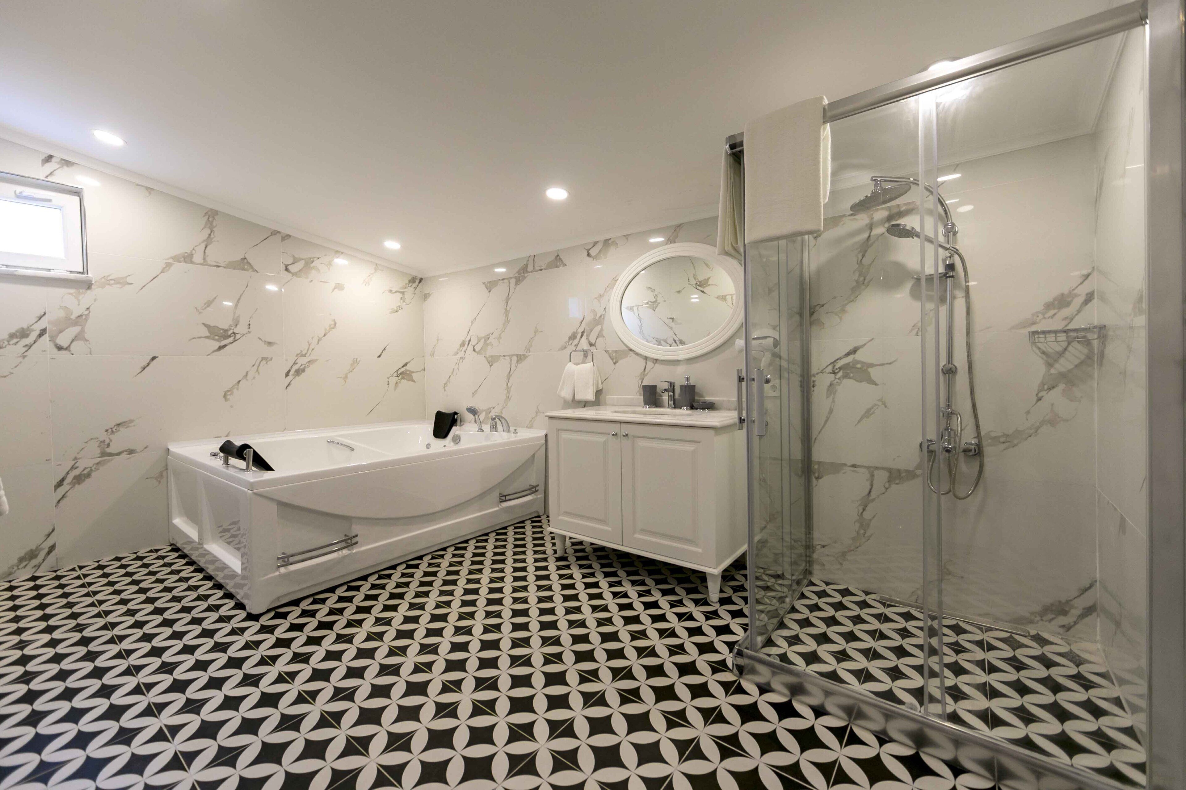 Un baño moderno con pared de mármol, piso a base de patrones en blanco y negro, una bañera independiente, ducha con cerramiento de vidrio y un espejo redondo sobre el lavamanos.
