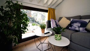 Una sala de estar con un sofá, una mesita redonda, una planta en maceta y una ventana con vistas a un paisaje montañoso.