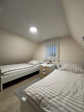 A bedroom with two single beds, a bedside table with a lamp, a window with sheer curtains, and a radiator.