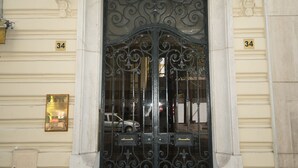 Ein Gebäudeeingang mit einem schwarzen, verzierten Tor, einem Balkon mit weißen Brüstungen und der Hausnummer 34 an der Wand.