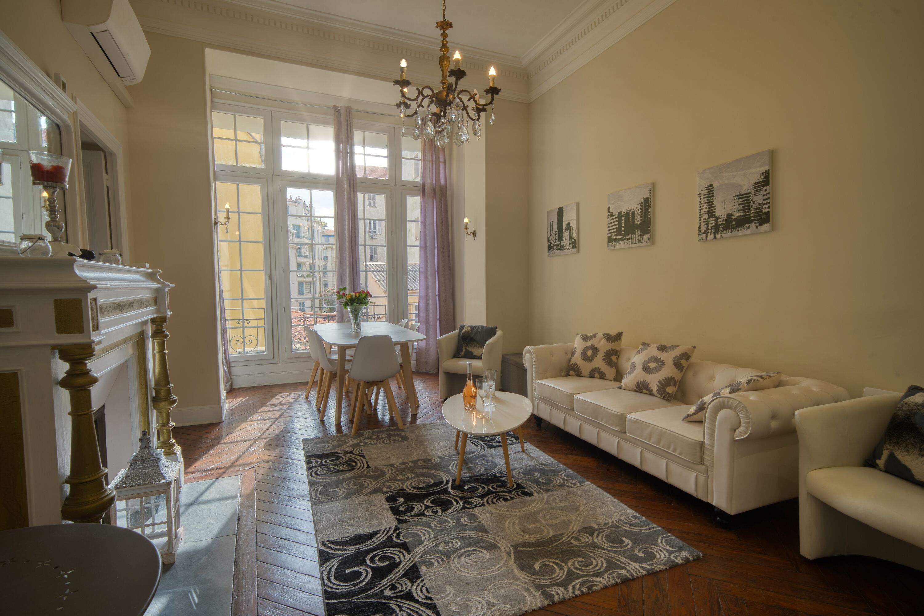 Een woonkamer met een witte bank, een kleine ronde tafel en een kroonluchter. Er is een groot raam met gordijnen, een open haard en een eethoek met een tafel en stoelen.