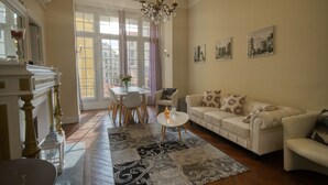 Ein Wohnzimmer mit einem weißen Sofa, einem kleinen runden Tisch und einer Kristallleuchter. Ein großes Fenster mit Vorhängen, ein Kamin und ein Essbereich mit Tisch und Stühlen sind ebenfalls vorhanden.