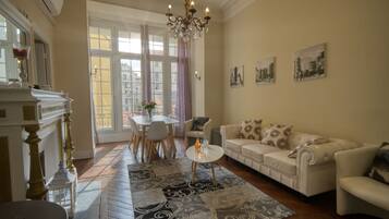 Een woonkamer met een witte bank, een kleine ronde tafel en een kroonluchter. Er is een groot raam met gordijnen, een open haard en een eethoek met een tafel en stoelen.