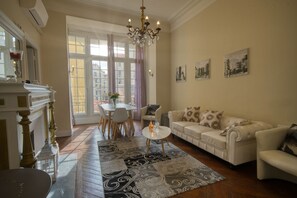 Ein Wohnzimmer mit einem weißen Sofa, einem kleinen runden Tisch und einer Kristallleuchter. Ein großes Fenster mit Vorhängen, ein Kamin und ein Essbereich mit Tisch und Stühlen sind ebenfalls vorhanden.