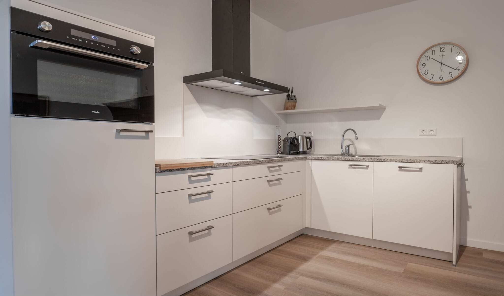 A modern kitchen with white cabinetry, a built-in oven, a stainless steel sink, and a wall clock.