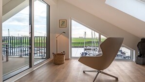 A modern loft bedroom with large windows overlooking a marina, a wooden floor, and a contemporary chair. - Breathtaking Penthouse Views (Kamperland)