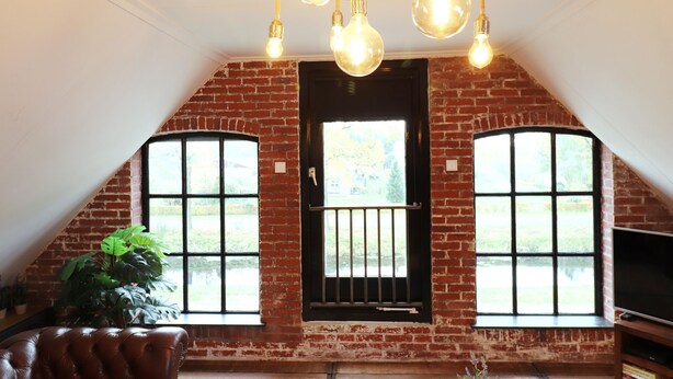 A room with exposed brick walls, a large window with a metal gate, and a ceiling with exposed beams and hanging light bulbs. - Shed 57b in an old flour mill in Haulerwijk (Haulerwijk)