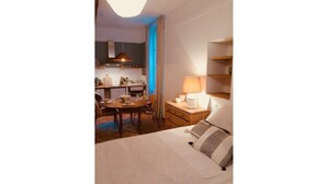 Camera d'albergo con un letto, una scrivania con lampada, un tavolo da pranzo con sedie e un angolo cottura con lavatrice.