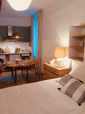 A hotel room with a bed, a desk with a lamp, a dining table with chairs, and a kitchen area with a washing machine.