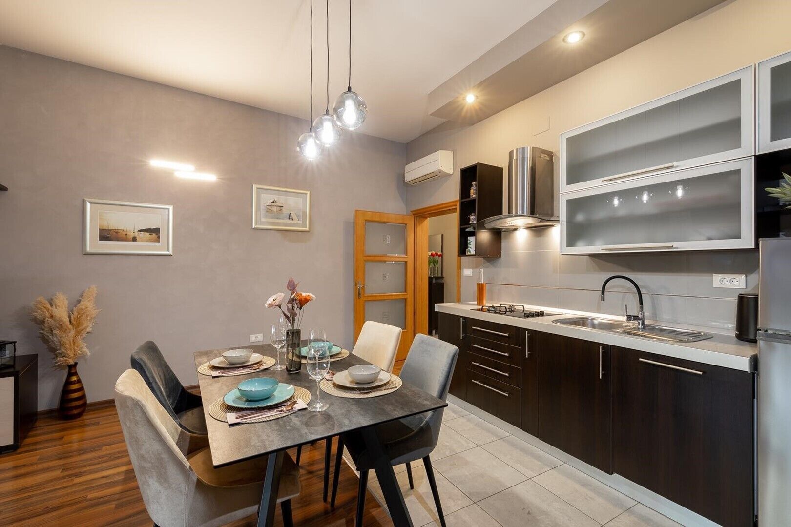 Una cocina moderna con armarios oscuros, un refrigerador de acero inoxidable y un horno empotrado. Hay una zona de comedor con una mesa dispuesta para dos personas, sillas y un florero con flores secas.