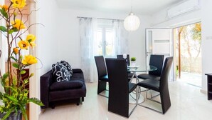 Ein modernes Wohnzimmer mit einem schwarzen Sofa, einem Glastisch und schwarzen Stühlen. Ein großes Fenster mit transparenten Vorhängen, eine Pendelleuchte und ein Topf mit gelben Blüten.