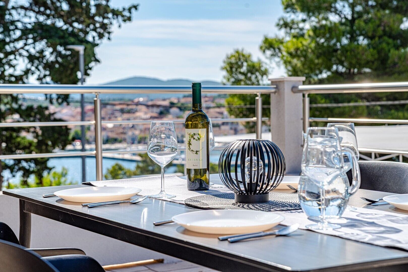 An outdoor dining table with a bottle of wine, glasses, and a decorative lamp.